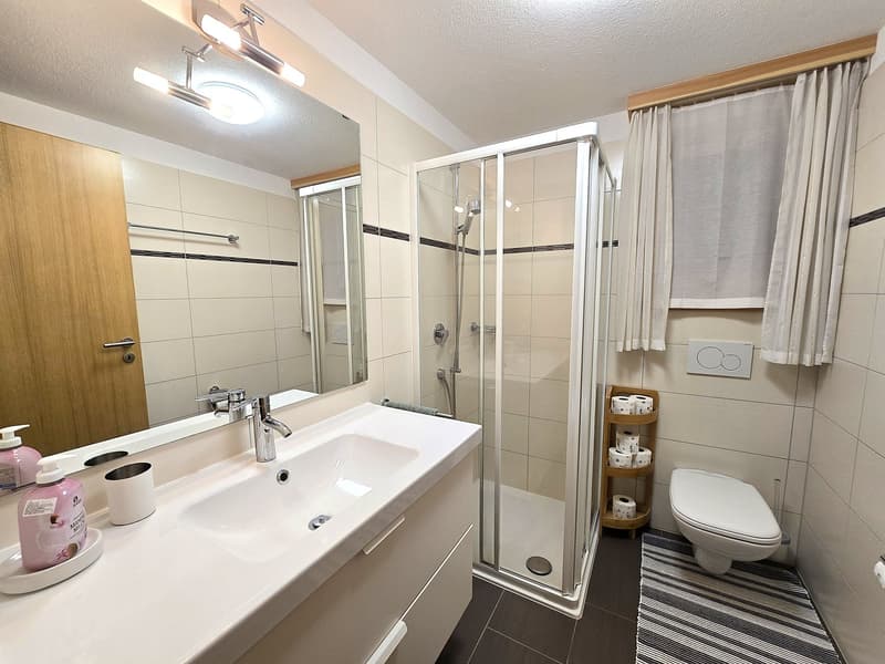 modern bathroom with white tiles, large mirror, glass shower, white sink, wooden cabinet, toilet, and toilet paper holder