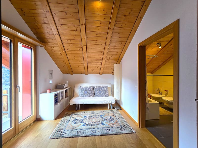 A cozy attic room with wooden ceiling, white walls, a white couch with pillows, a white shelf, a rug, and a glass door leading to a balcony. The room also has a white toilet and sink in the bathroom.