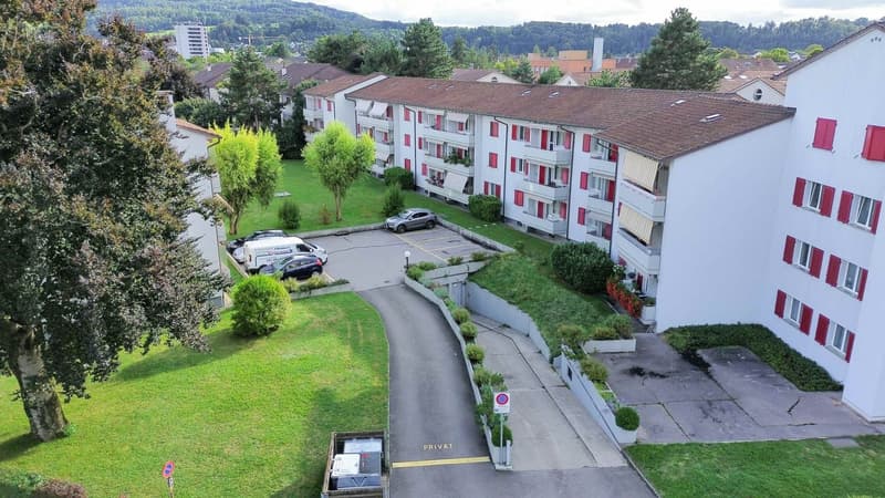 Three-story residential building, red shutters, parking area in front, car parking spaces marked, some cars parked