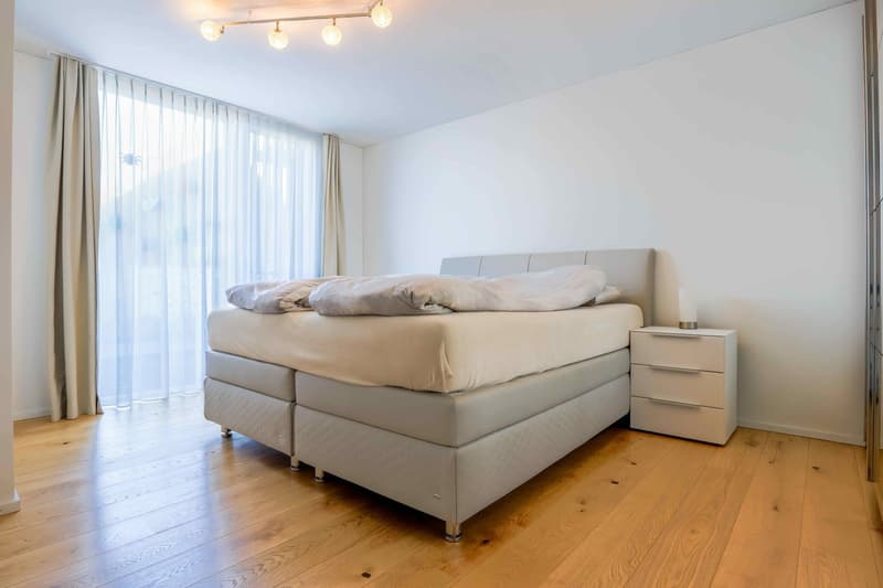 Double bed with headboard, side table, white nightstand, white wall, wooden floor, large window with curtains, modern lighting