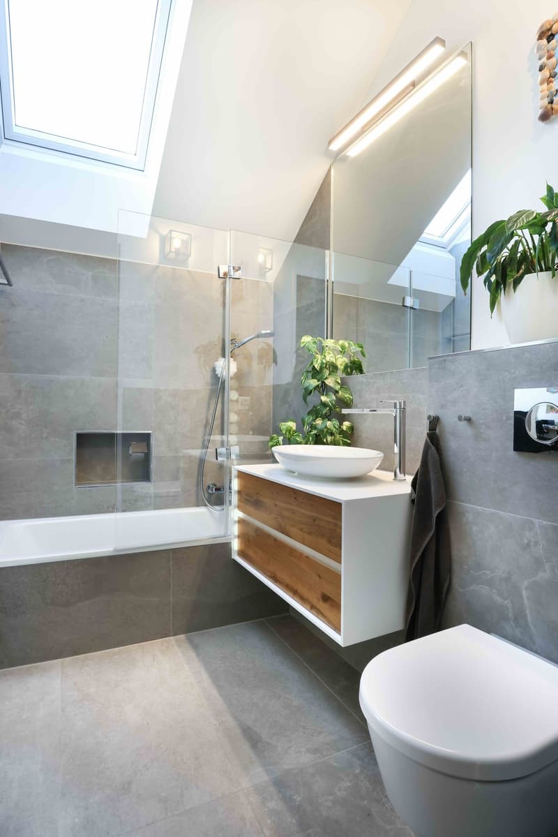 Bathtub, white sink, wooden cabinet, gray tiles, skylight window, modern fixtures, showerhead, faucet, potted plant