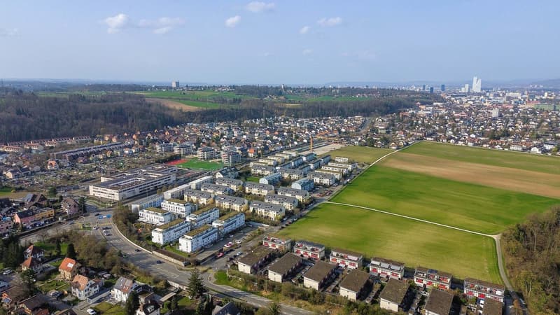 Aerial view of a residential area with multiple buildings, green fields, and a road.