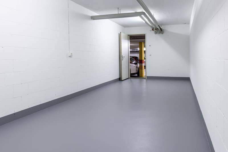 Empty room with white walls, gray floor, open door to another room with a car parked