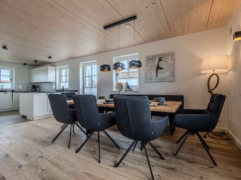 Dining room with wooden floor, wooden dining table, modern chairs, pendant lights, white cabinets, kitchen area, windows, artwork, lamp
