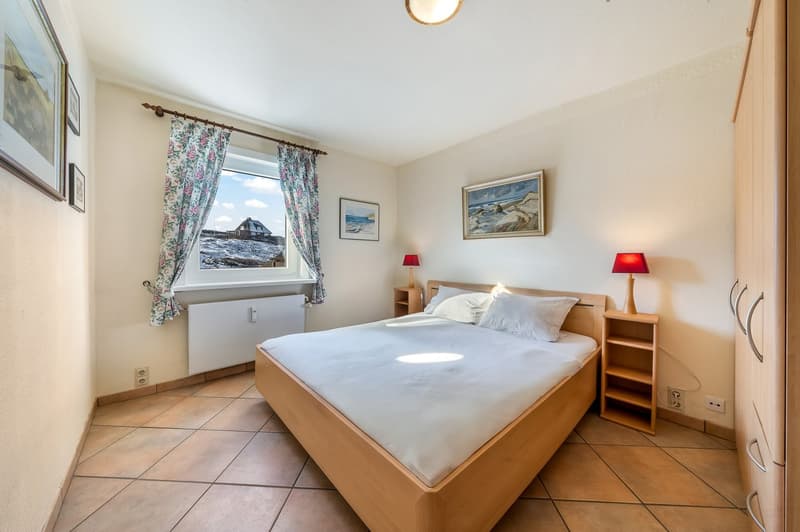 Bedroom with bed, nightstands, lamps, window with curtains, radiator, picture frames, tiled floor, wardrobe