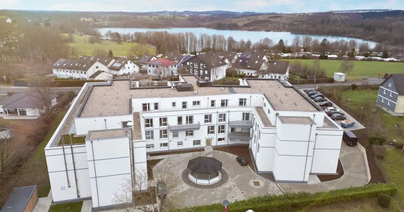 Aerial view of a modern white building with multiple windows, a flat roof, and a small gazebo in the center. The building is surrounded by a paved area and greenery. Several cars are parked in front of the building. In the background, there is a lake and