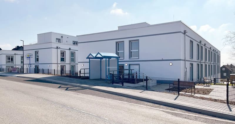 Modern white building, multiple windows, entrance with canopy, metal fence, paved sidewalk, street light