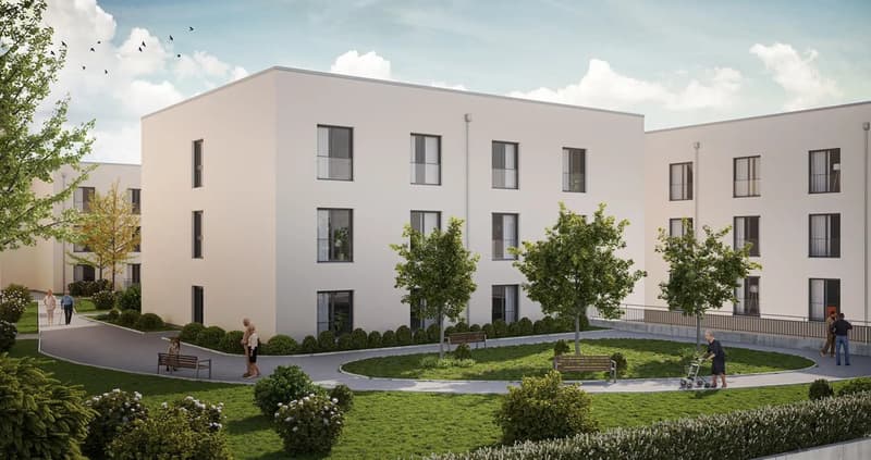 A modern white building with multiple windows, surrounded by a well-maintained garden with trees, bushes, and a paved walkway. There are benches and a person with a trolley in the garden. The building has a flat roof and is situated in a peaceful environm