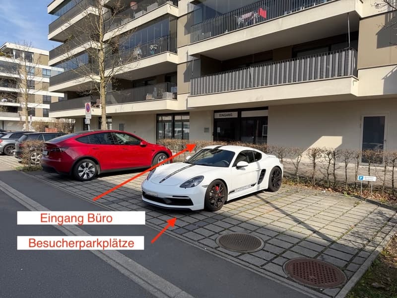 A multi-story building with glass windows and balconies. Two cars are parked in front of the building, one is a red Tesla and the other is a white Porsche. The building has a sign that reads 'Eingang' and 'Besucherparkplatz'. There are bushes and a tree i