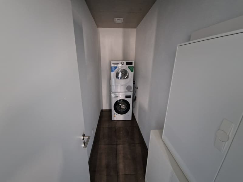 A small room with a white washing machine and a white dryer stacked on top of each other, white walls, and brown tiled floor.