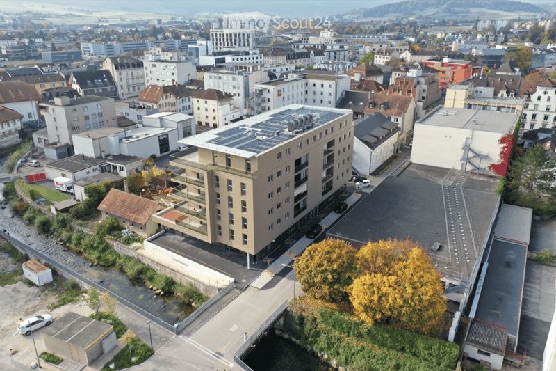 Aerial view of a multi-story building with solar panels on the roof, surrounded by other buildings and greenery.