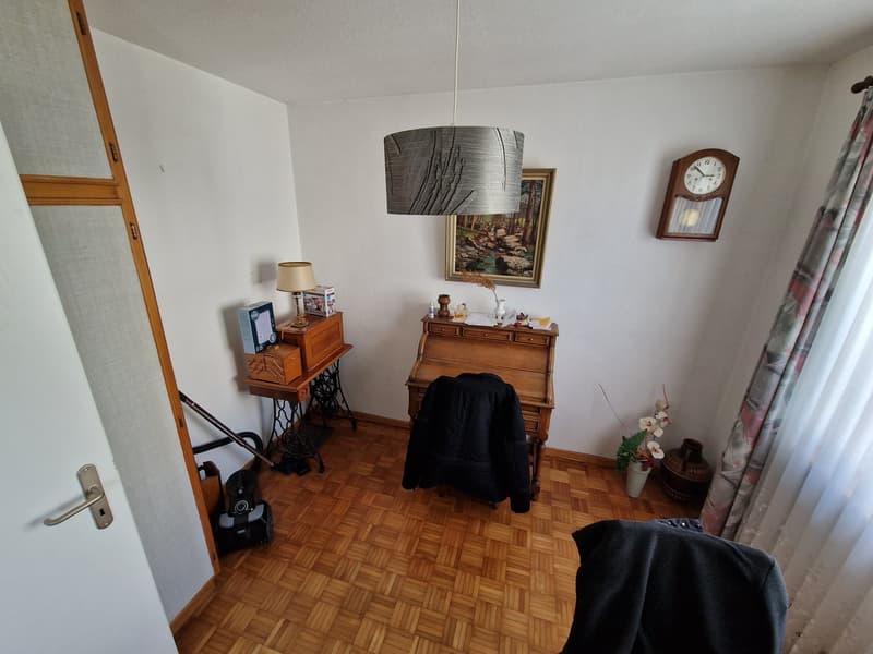 wooden floor, white walls, wooden desk, sewing machine, wooden cabinet, lamp, clock, picture frame, flowers, jacket
