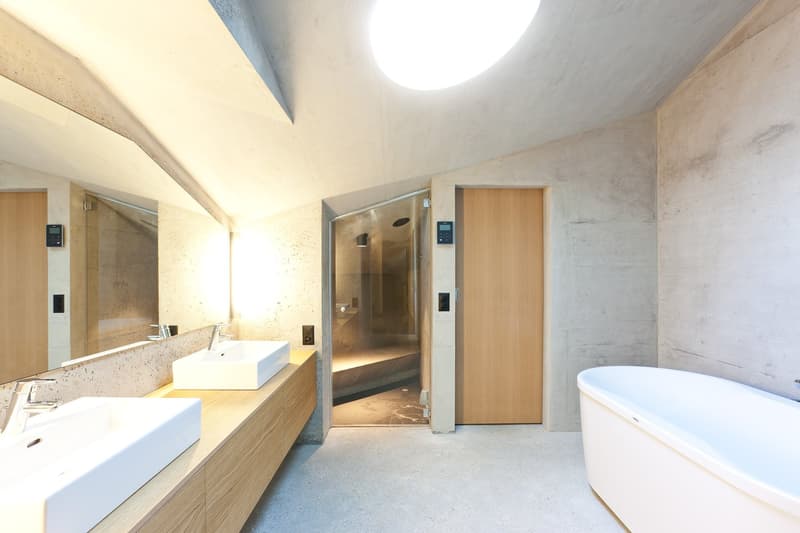 modern bathroom with concrete walls, wooden door, large mirror, two sinks, bathtub, glass shower