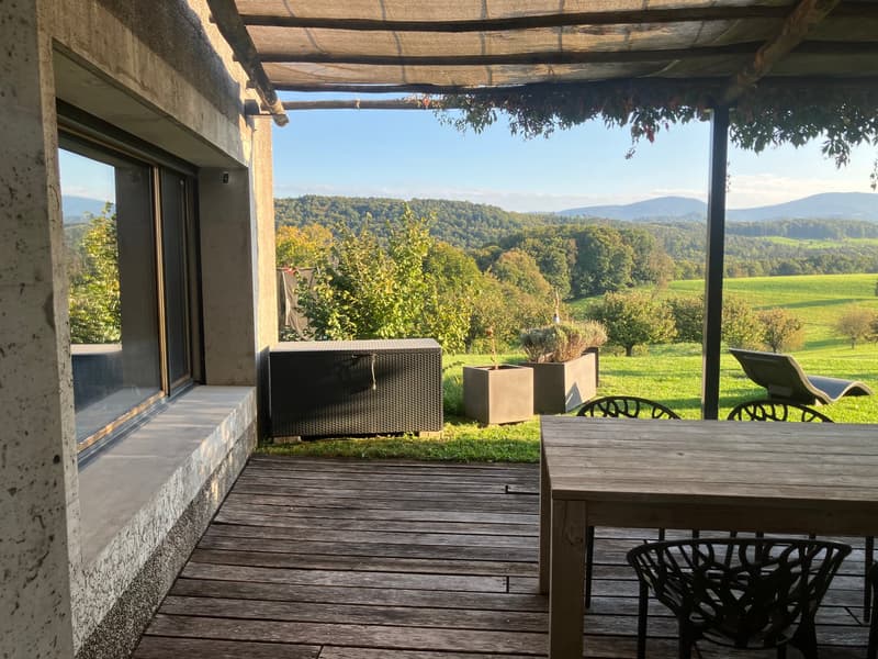 Wooden deck with a table and chairs, covered by a pergola, overlooking a grassy area with plants and a distant view of mountains