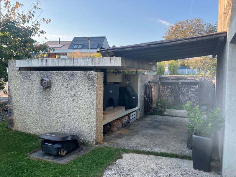 Outdoor kitchen with a stone wall, a wooden canopy, a black oven, a black cover, a wooden shelf, a black lawn mower, a potted plant, and a grassy area.