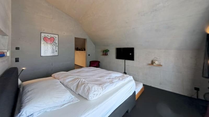 bedroom with a double bed, white bedding, concrete walls, mounted TV, artwork, small table, plant, wooden cabinet, chair