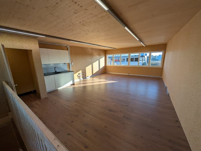 Empty room with wooden floor, white cabinets, kitchen area with sink, large windows, wooden railing