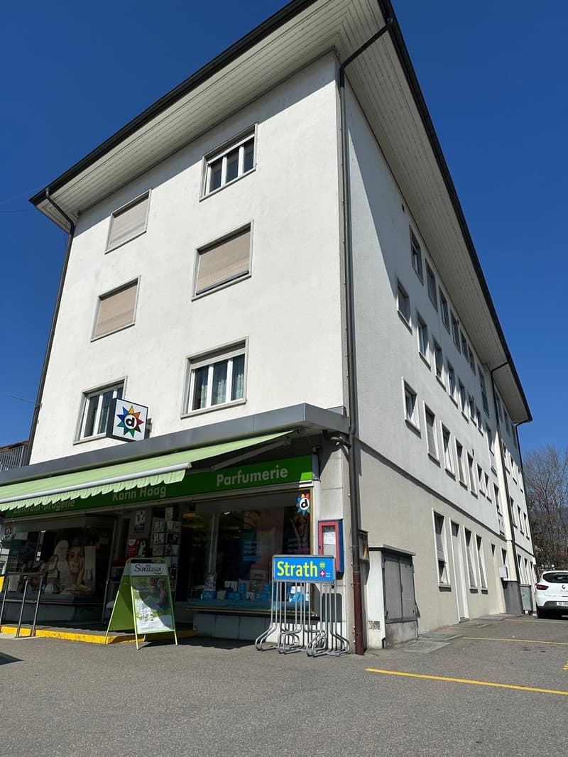 A large white building with multiple floors, a shop on the ground floor with a green awning, and a car parked in front.
