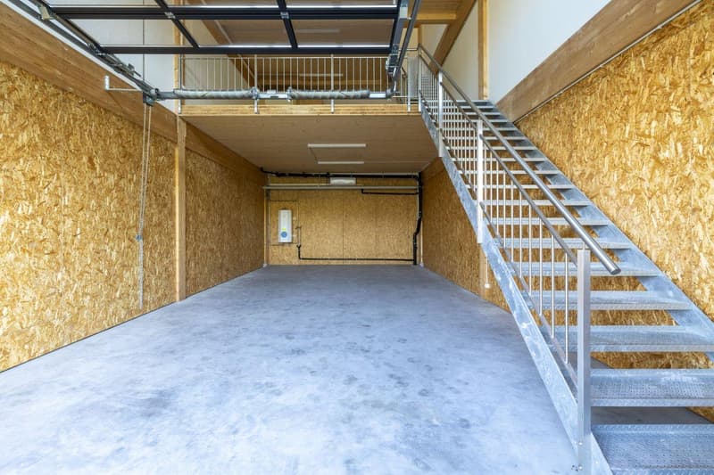 The image shows a large, open garage space with a concrete floor and a metal staircase leading to an upper level. The walls are made of oriented strand board (OSB) panels, and there are various pipes and ducts running along the ceiling.
