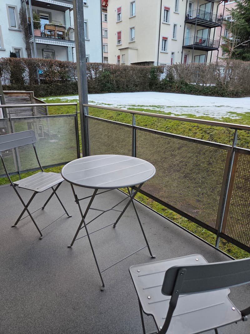 Two metal folding chairs and a small table on a balcony with a metal railing, a grassy field and a building in the background