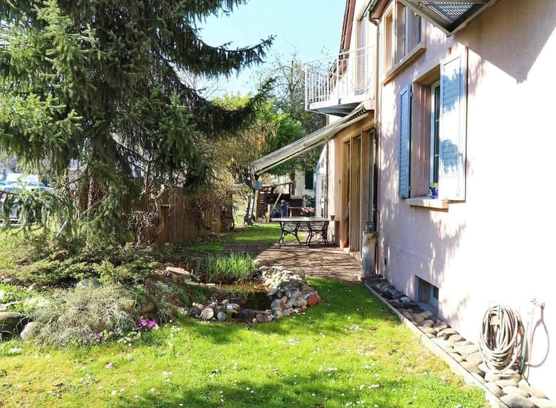 House backyard with balcony, garden with plants, small pond, outdoor table and bench, and fence