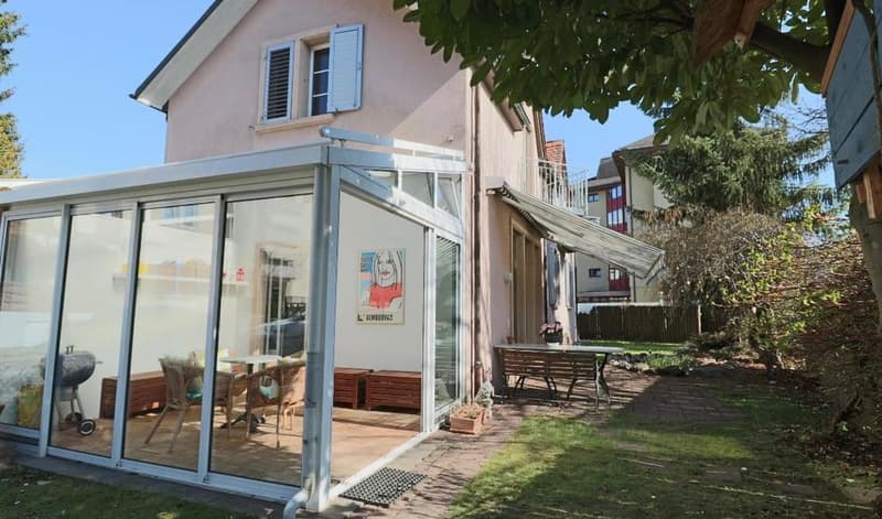House with pink exterior, glass walls, balcony, table and chairs, and a potted plant