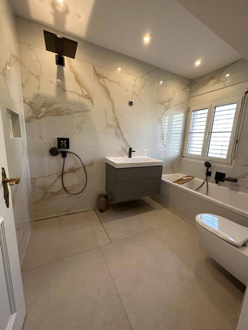 Modern bathroom with marble tiles, walk-in shower with rain shower head, freestanding bathtub, white vanity with sink and drawers, and white toilet. Floor is tiled and there is a trash can and small tray by the bathtub.