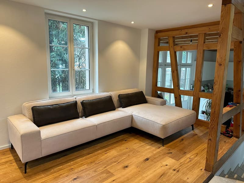 A corner sofa, wooden floor, white walls, window with wooden frames, an open space connected to the living area.