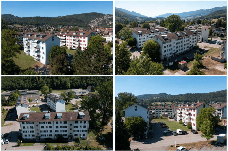 Aerial view of a residential building complex with multiple buildings, surrounded by trees and greenery, with a parking area and a road in front.