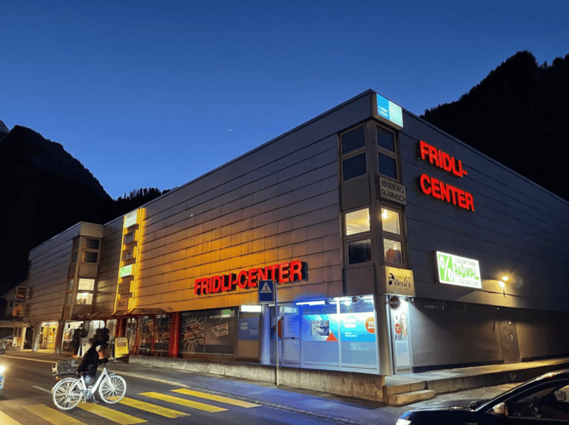 A large building with a sign that reads 'FRIDLI-CENTER', multiple windows, and a person riding a bicycle in front.