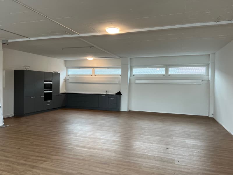 An empty kitchen with hardwood flooring, white walls, ceiling lights, modern cabinetry, integrated oven and microwave, sink with faucet, and large windows.