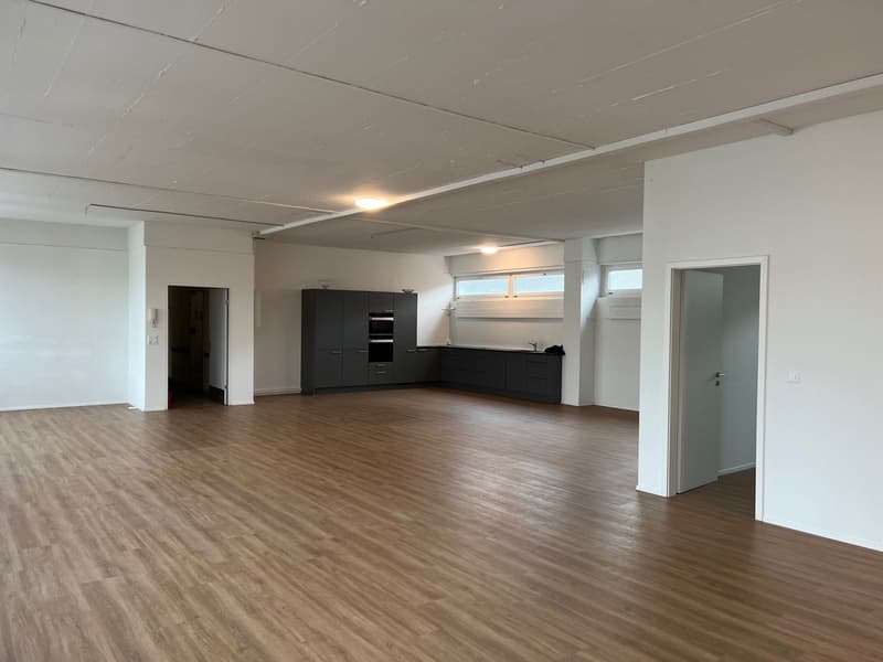 Empty room with hardwood floors, white walls, built-in cabinets, open kitchen area, windows, ceiling lights