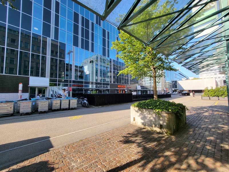 Large glass-walled commercial building with a covered walkway, trees, and a Migros supermarket visible. The building has a modern, sleek design with a mix of glass and metal materials.
