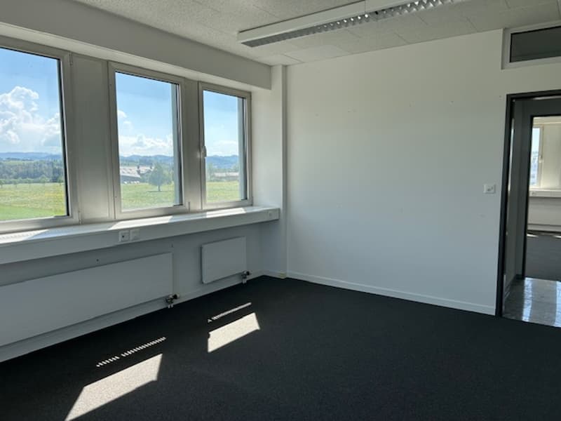 Spacious office space with large windows providing a scenic view of the surrounding landscape. The room has white walls, a black carpet floor, and appears to be a newly renovated or unoccupied space.