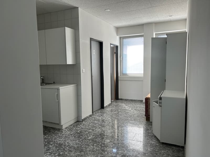 The image shows a spacious apartment interior with a kitchen area, a hallway, and multiple doors leading to other rooms. The floor is covered in a gray granite-like tile, and the walls are painted in a light, neutral color. The kitchen area has built-in c