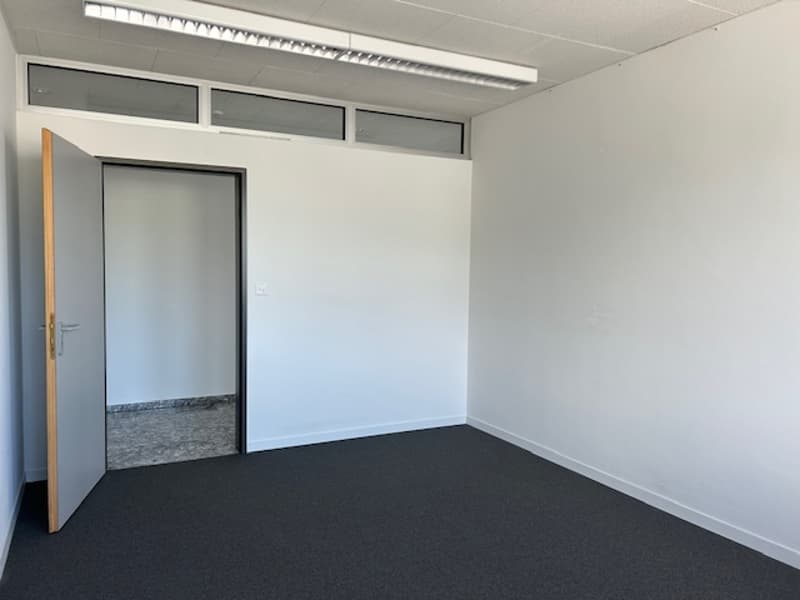 The image shows a simple, minimalist office space with white walls, a black floor, and a glass door. The room appears to be well-lit with windows near the ceiling.