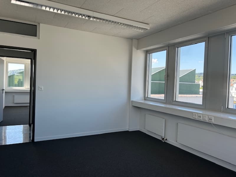 The image shows a spacious office space with large windows, white walls, and a black carpet floor. The room appears to be empty, with no furniture or other objects visible.