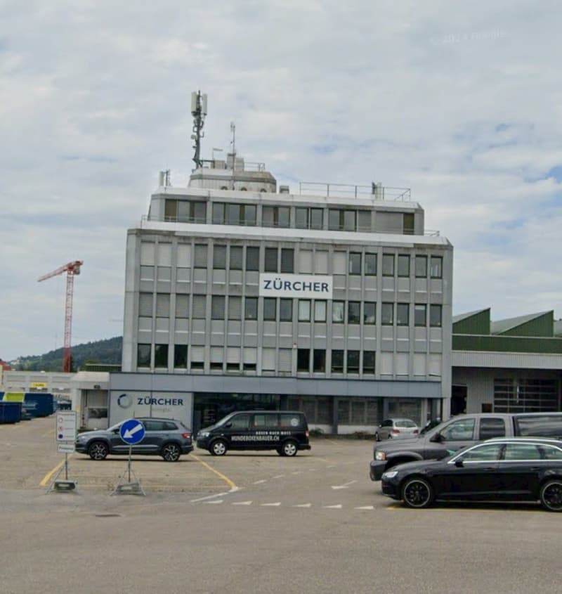 Multi-story office building with the Zurcher logo prominently displayed, surrounded by parked cars in a parking lot.