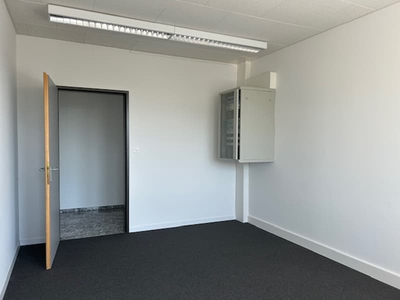 The image shows a simple, minimalist office space. The room has white walls, a black floor, and a door on the left side. There is a utility cabinet or electrical panel mounted on the wall. The lighting fixtures on the ceiling suggest this is a commercial