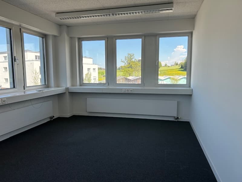 The image shows a spacious office space with large windows providing a view of the outdoor scenery. The room has white walls, a black carpeted floor, and appears to be empty, ready for use.