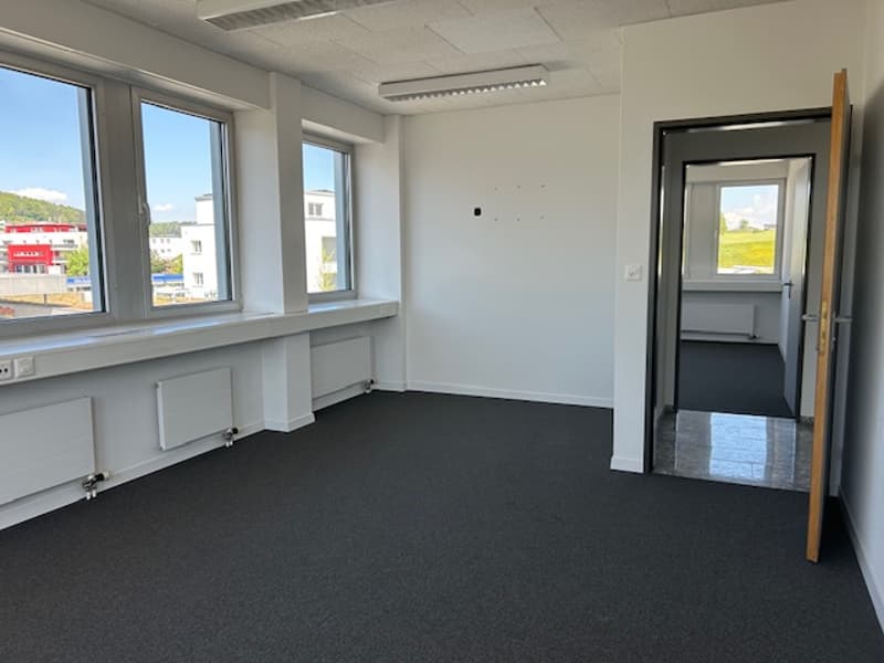 The image shows a spacious office space with large windows, white walls, and a black carpeted floor. The room appears to be empty, with no furniture or decor visible. There is a doorway leading to another room or area visible in the background.