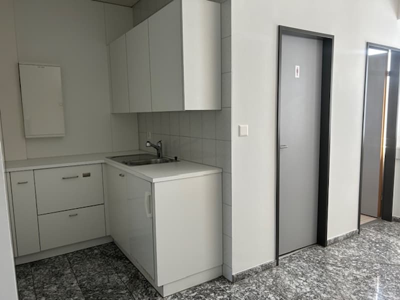 The image shows a modern, minimalist kitchen with white cabinets, a stainless steel sink, and a dishwasher. The floor is covered in gray tile, and there is a doorway leading to another room.