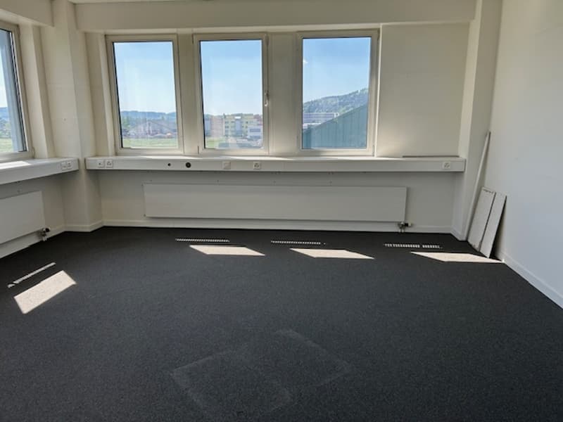 Large open office space with floor-to-ceiling windows providing a scenic view of the surrounding area. The room has a dark gray carpeted floor and white walls.