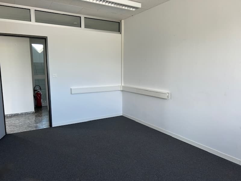 The image shows an empty office space with white walls, a gray floor, and a window. The room appears to be a simple, minimalist office space.
