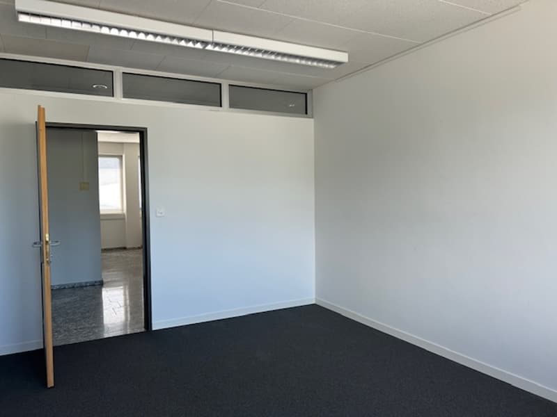 The image shows an empty office space with white walls, a gray floor, and a door leading to another room. The room has overhead lighting and appears to be a clean, modern office environment.