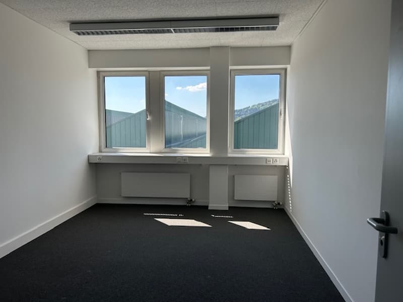 Empty office space with large windows, white walls, and black carpeted floor.