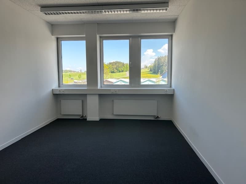 The image shows an empty office space with large windows overlooking a scenic outdoor landscape. The room has gray walls, a black carpeted floor, and built-in desks or workstations along the walls.