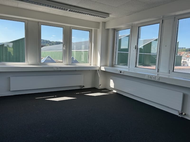 Large open office space with multiple windows providing a view of the surrounding area, grey walls, black carpeted floor, and electrical outlets along the walls.