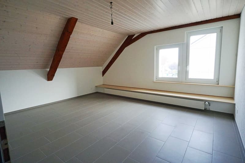 Attic space with slanted ceiling, wooden beams, and large windows. The room has a tiled floor and a built-in bench or seating area along one wall.