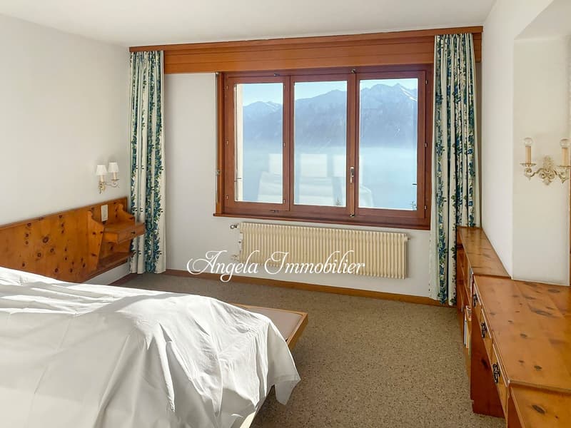 bedroom with bed, wooden headboard, wooden shelves, large window, curtains, radiator, wooden floor, white walls